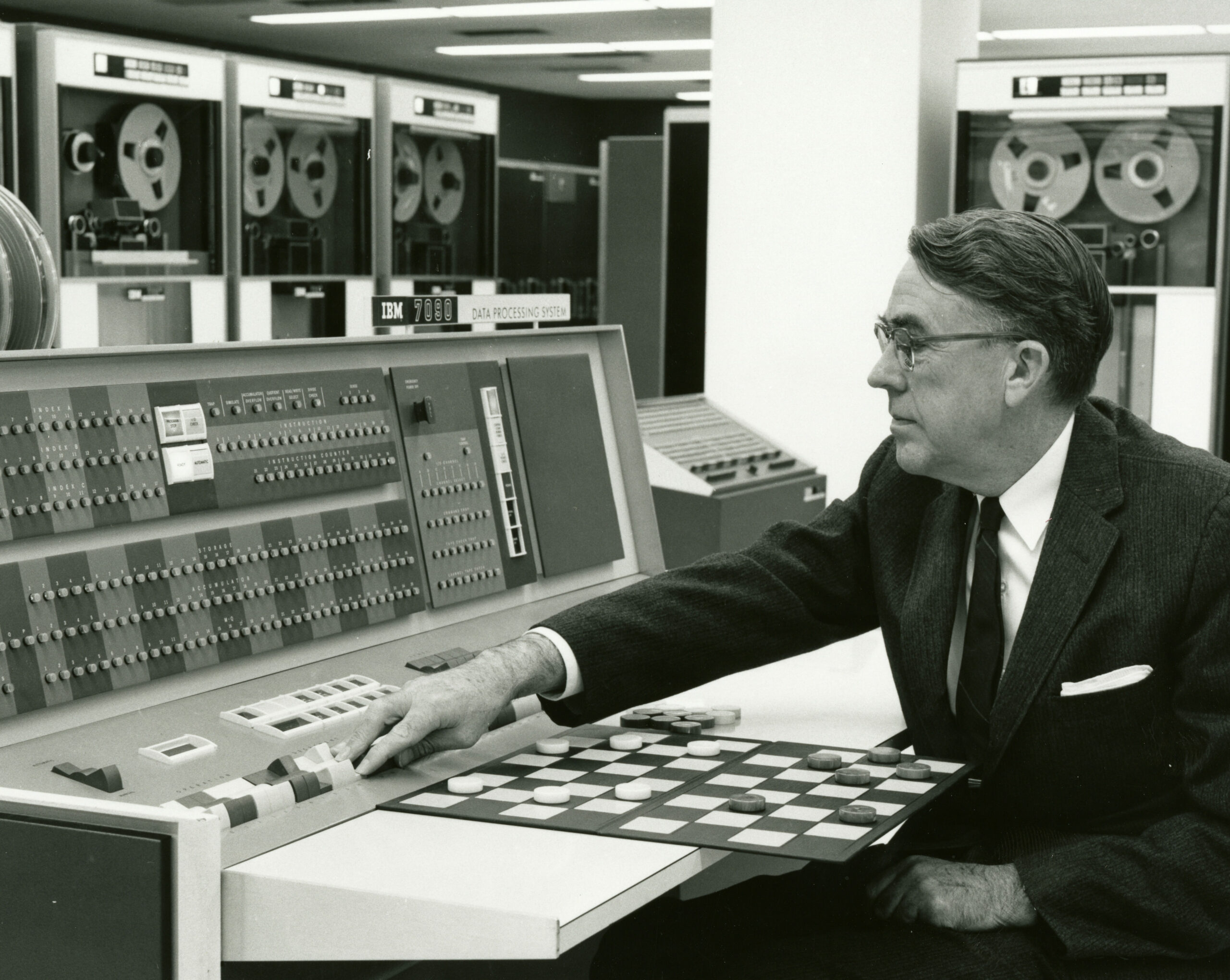 Schwarz-weiß Fotografie aus den 60ern. Rechts im Bild sitzt ein Mann im Anzug der eine Maschine mit vielen Knöpfen bedient, Dabei greift er über ein Spielbrett des Spiels Dame. Englische Bildunterschrift:  Arthur Samuel demonstrates how machine learning can be used to play checkers in 1962