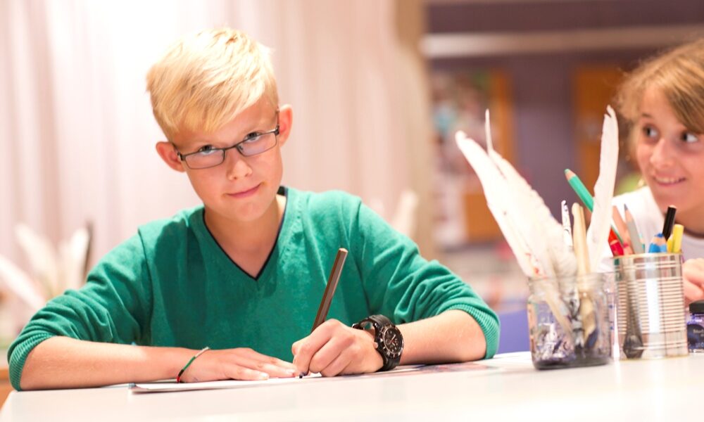 Ein Kind sitzt am Tisch und schreibt mit einem braunen Buntstift etwas auf ein Blatt Papier. Neben dem Kind ist ein Glas mit Schreibfedern und eine Dose mit Stiften.