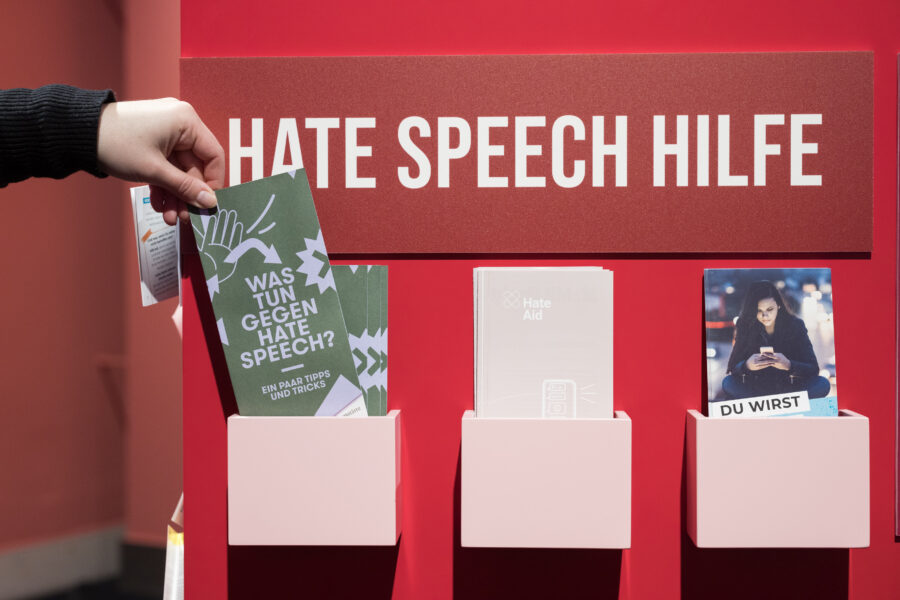Eine rote Wand auf der in weißer Schrift Hate Speech Hilfe steht. Unter derm Schriftzug sind drei unterschiedliche Flyer. Ganz links greift eine Hand nach einem Flyer auf dem steht Was tun gegen Hate Speech?