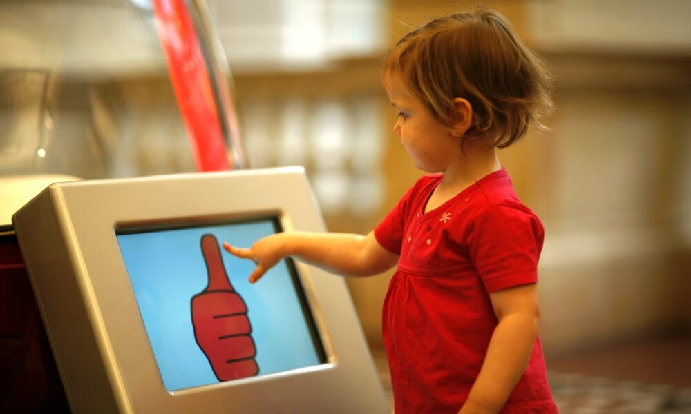 Ein Kind steht rechts neben einem Bildschirm und tippt mit einem Finger auf den Bildschirm. Auf dem Bildschirm ist auf weißem Grund ein roter Daumen nach oben abgebildet.