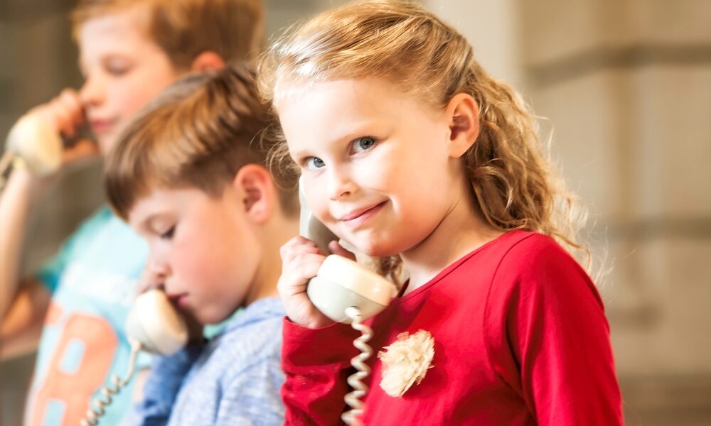 Im Vordergrund ist ein Mädchen mit einem Telefonhörer den sie sich ans ohr hält. Im Hintergrund sind zwei weitere Kinder die sich einen Telefonhörer ans Ohr halten.